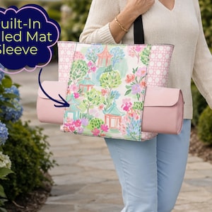 May include: A tote bag featuring a central panel with a colorful garden print of pagodas and trees, flanked by pink side panels with a white circular pattern. The bag includes a built-in rolled mat sleeve containing a solid pink rolled mat.