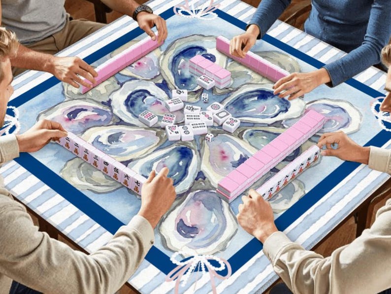 May include: A group of people playing Mahjong. The game is set on a table with a blue and white oyster-themed design. Pink tile racks and white tiles are visible, with players' hands around the table.