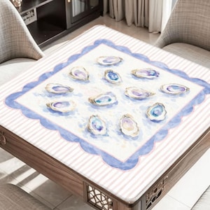 May include: A wooden table with a white and pink striped surface. The table has a square, scalloped blue border with watercolor oyster shell illustrations. The table is surrounded by two chairs.