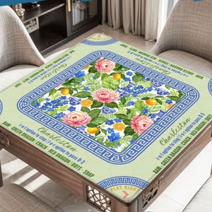 May include: A square table with a floral design, featuring pink roses, blue flowers, and orange fruit. The table has a light green border with a blue Greek key pattern and text that reads "Charleston". The table is made of wood.