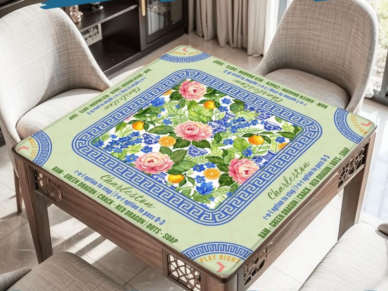 May include: A square table with a floral design, featuring pink roses, blue flowers, and orange fruit. The table has a light green border with a blue Greek key pattern and text that reads "Charleston". The table is made of wood.