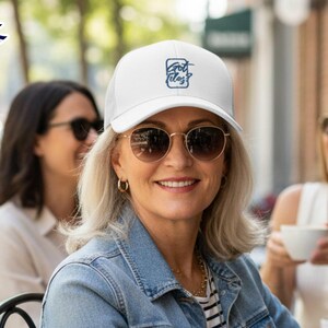 Op de afbeelding: Een witte baseballcap met een mesh rug en de tekst "Got Ideas?" in een blauw vierkant. De persoon draagt een spijkerjasje en een zonnebril met gouden montuur. Twee andere mensen zijn op de achtergrond.