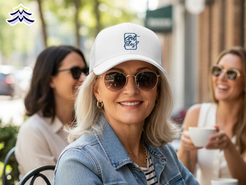 Op de afbeelding: Een witte baseballcap met een mesh rug en de tekst "Got Ideas?" in een blauw vierkant. De persoon draagt een spijkerjasje en een zonnebril met gouden montuur. Twee andere mensen zijn op de achtergrond.