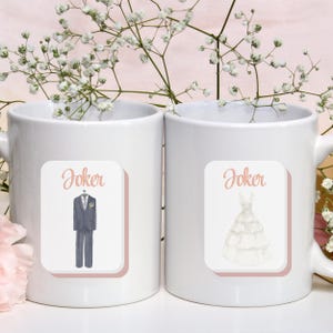 May include: Two white ceramic mugs with the word "Joker" above illustrations. One mug features a watercolor image of a gray suit and the other a white wedding dress. The mugs are decorated with a pink border and are surrounded by white flowers.