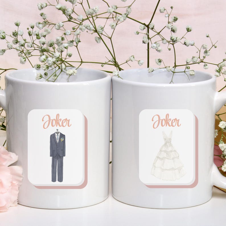 May include: Two white ceramic mugs with the word "Joker" above illustrations. One mug features a watercolor image of a gray suit and the other a white wedding dress. The mugs are decorated with a pink border and are surrounded by white flowers.