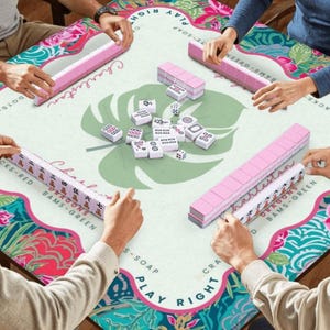 May include: A group of people playing Mahjong on a table with a floral design. The table has a light green background with a large leaf design in the center. Pink tiles and racks are visible, with the word "Charleston" printed on the table.