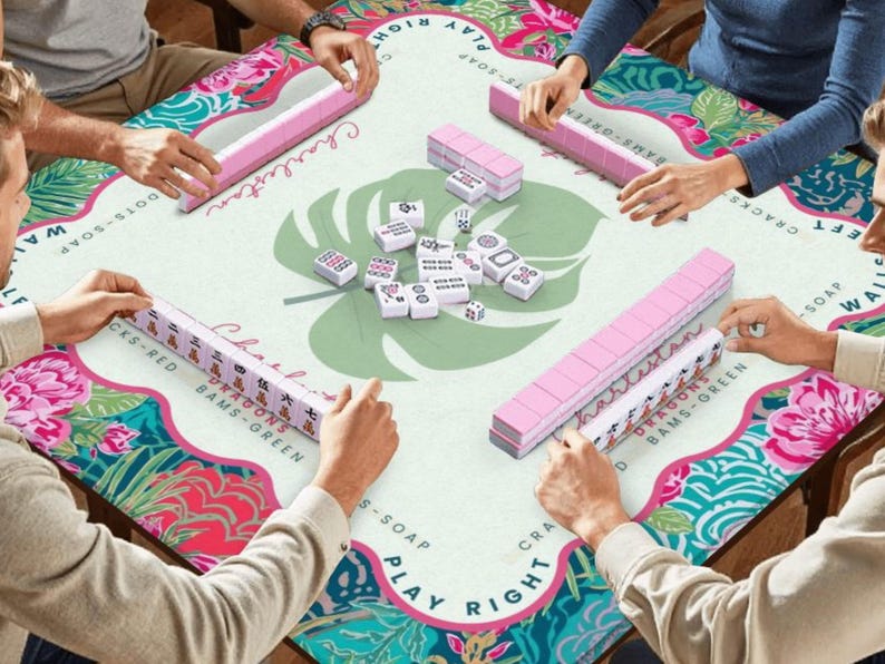 May include: A group of people playing Mahjong on a table with a floral design. The table has a light green background with a large leaf design in the center. Pink tiles and racks are visible, with the word "Charleston" printed on the table.