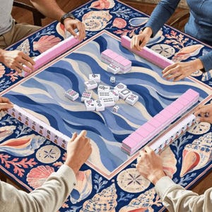 K&ouml;nnte beinhalten: Mahjong-Spiel in Aktion, mit Spielern um einen Tisch. Der Tisch hat ein zentrales blau-wei&szlig;es Wellenmuster, umrahmt von einem Muschelrand. Rosa Spielsteine und Halterungen sind zu sehen.