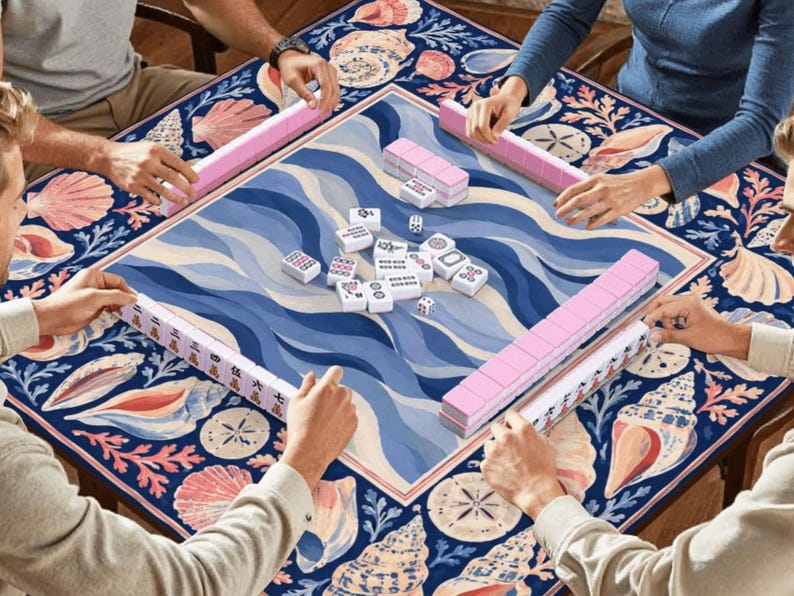 K&ouml;nnte beinhalten: Mahjong-Spiel in Aktion, mit Spielern um einen Tisch. Der Tisch hat ein zentrales blau-wei&szlig;es Wellenmuster, umrahmt von einem Muschelrand. Rosa Spielsteine und Halterungen sind zu sehen.