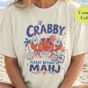 May include: A cream-coloured short-sleeve t-shirt featuring a graphic of a red crab wearing heart-shaped sunglasses. The text reads If CRABBY PLEASE RETURN TO MAHJ. The design includes mahjong tiles, a palm tree, a seashell, and a starfish.