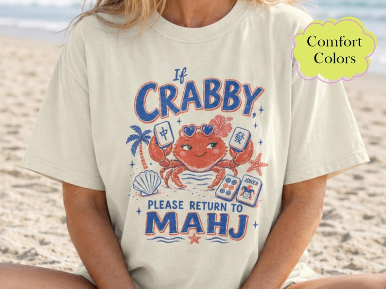 May include: A cream-coloured short-sleeve t-shirt featuring a graphic of a red crab wearing heart-shaped sunglasses. The text reads If CRABBY PLEASE RETURN TO MAHJ. The design includes mahjong tiles, a palm tree, a seashell, and a starfish.