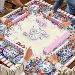 May include: A square mahjong table covered with a festive tablecloth featuring blue and white floral patterns, red ribbons, and holly berries. Four players are seated around the table, each with a rack of pink tiles, while white tiles are in the centre.