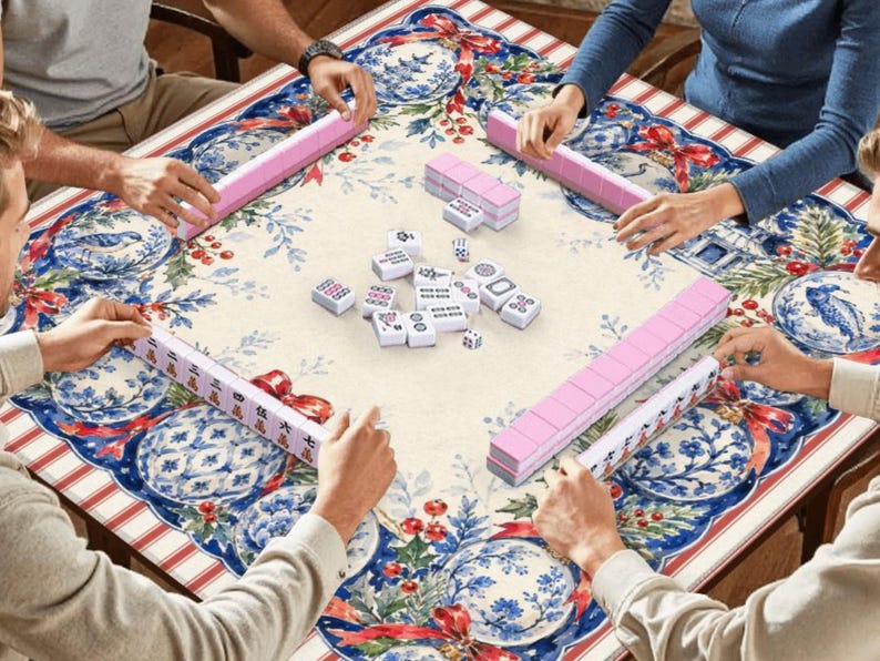 May include: A square mahjong table covered with a festive tablecloth featuring blue and white floral patterns, red ribbons, and holly berries. Four players are seated around the table, each with a rack of pink tiles, while white tiles are in the centre.