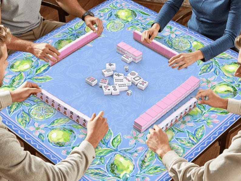 May include: A group of people playing Mahjong. The game is set on a blue table with a floral and citrus fruit design. Pink Mahjong tiles are arranged around the table, with white tiles and dice in the center.