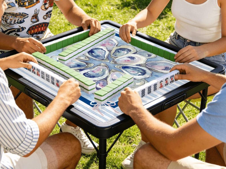 May include: A square Mahjong table with a black frame and a blue and white oyster shell design. Green and white tiles are arranged around the table as people play the game outdoors on a sunny day.