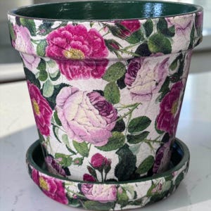 May include: A tapered ceramic flower pot with a matching saucer, featuring a detailed floral pattern of vibrant magenta and soft pink roses with green leaves on a white background, finished with a solid dark green interior and rim.