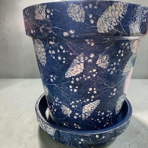 May include: A tapered cylindrical plant pot with a matching saucer, featuring a deep navy blue base color decorated with a repeating white pattern of pinecones, thin branches, and small berries, finished with a glossy, textured surface.