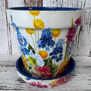 May include: A white ceramic flower pot with a matching saucer, both adorned with a vibrant floral design. The pot features a textured surface and a dark blue rim. The colorful flowers include yellow, blue, and pink blooms.