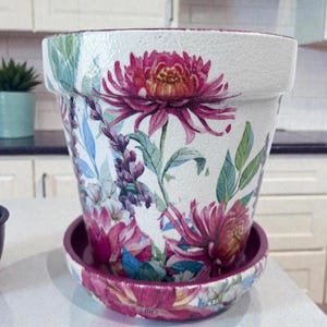 May include: A white ceramic flower pot with a matching saucer, decorated with a floral design. The design features large, pink and purple flowers with green leaves and stems. The pot is approximately 7 inches tall and 8 inches in diameter.