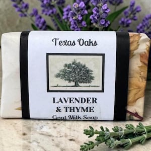May include: A rectangular bar of Texas Oaks Lavender & Thyme goat milk soap wrapped in paper with a central white label featuring a black and white illustration of an oak tree, black text, and a decorative leaf pattern on the side edges.