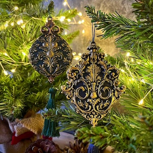 May include: Two hanging ornaments featuring intricate, raised gold-toned scrollwork patterns against a matte black base. Each ornament has a decorative metal top loop and a long, dangling fabric tassel in dark green or navy blue.