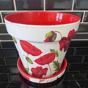 May include: A white ceramic flower pot with a red interior and matching saucer. The pot is decorated with red poppy flower illustrations and green leaves. The pot is approximately 7 inches in diameter.
