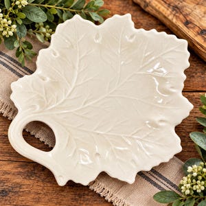 May include: A cream-colored ceramic serving platter shaped like a large maple leaf. The platter features a glossy finish, embossed vein details across the surface, and a circular cutout handle integrated into the stem area of the leaf design.