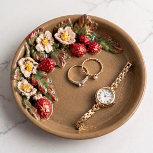 May include: A round, tan ceramic trinket dish featuring a raised, three-dimensional relief of white flowers, green leaves, and red strawberries along one edge. Inside the dish are two gold-toned rings and a gold-toned wristwatch with a white face.