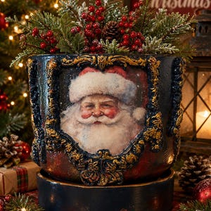 May include: A cylindrical dark blue and red planter featuring a central portrait of Santa Claus framed by ornate gold-coloured relief detailing. The planter is filled with artificial pine branches, red berries, and a small pinecone.