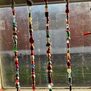 Op de afbeelding: Decoratieve kralen raamhangers. Elke streng heeft een verscheidenheid aan kleurrijke kralen in rood, blauw, groen en bruin. De kralen zijn gemaakt van verschillende materialen en vormen, waardoor een unieke en artistieke uitstraling ontstaat. De strengen hangen voor een raam.