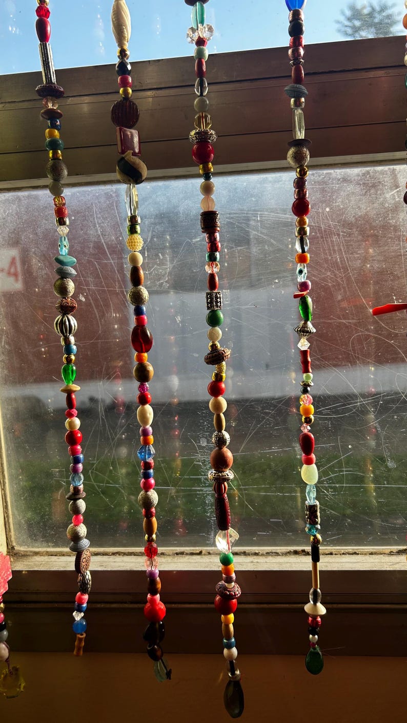 Op de afbeelding: Decoratieve kralen raamhangers. Elke streng heeft een verscheidenheid aan kleurrijke kralen in rood, blauw, groen en bruin. De kralen zijn gemaakt van verschillende materialen en vormen, waardoor een unieke en artistieke uitstraling ontstaat. De strengen hangen voor een raam.