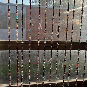 Op de afbeelding: Een kralengordijn hangt voor een raam. De strengen bestaan uit kleurrijke kralen in verschillende vormen en maten, waaronder rood, bruin, groen en blauw. Een decoratief element.