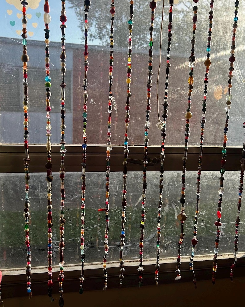 Op de afbeelding: Een kralengordijn hangt voor een raam. De strengen bestaan uit kleurrijke kralen in verschillende vormen en maten, waaronder rood, bruin, groen en blauw. Een decoratief element.