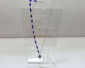Op de afbeelding: Een delicate ketting met een zilveren ketting en een hanger. De ketting heeft een streng blauwe en witte kralen. De hanger is een kleine, donkergrijze hartvorm. De ketting wordt tentoongesteld op een heldere acryl standaard.