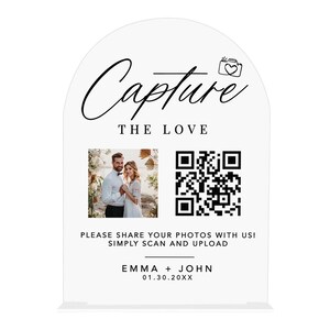 May include: A white acrylic wedding sign with an arched top. The sign features the words "Capture The Love" in elegant script, a photo of a couple, a QR code, and the names "Emma + John" with a date. The sign encourages guests to share photos.