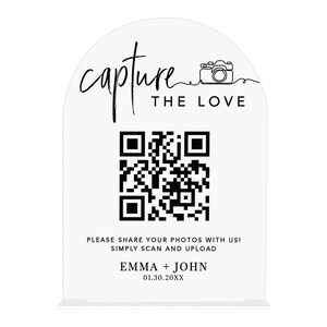 May include: Clear acrylic wedding sign with a curved top. The sign features the words "capture the love" and a QR code. Below the QR code is the text "Please share your photos with us! Simply scan and upload" and the names "EMMA + JOHN" with a date.