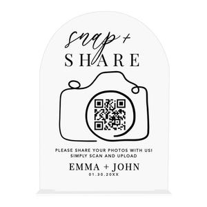 May include: A white acrylic sign with black text that reads "snap + SHARE" above a camera graphic with a QR code. Below the camera, it says "PLEASE SHARE YOUR PHOTOS WITH US! SIMPLY SCAN AND UPLOAD" and "EMMA + JOHN 01.30.20XX".