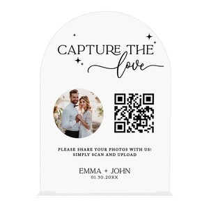 May include: A white wedding sign with the text "CAPTURE THE love" and a photo of a couple. Includes a QR code and the names "EMMA + JOHN" with a date. The sign is arched at the top.