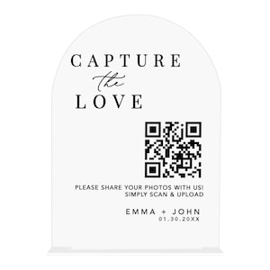 May include: A white acrylic wedding sign with an arched top. The sign reads "CAPTURE the LOVE" above a QR code and the text "PLEASE SHARE YOUR PHOTOS WITH US! SIMPLY SCAN & UPLOAD". The names "EMMA + JOHN" and a date are at the bottom.