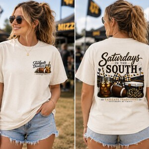 May include: A cream-coloured short-sleeve t-shirt featuring a small graphic on the front left chest with a football and drink, and a large back graphic with the text "Saturdays in the South" above a football, a drink, a bow, and a cooler.