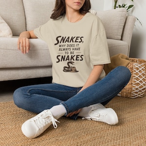 May include: Beige t-shirt with the text "Snakes, why does it always have to be snakes" in dark brown. The shirt features a graphic of a snake on a hat. The person is wearing blue jeans and white trainers.