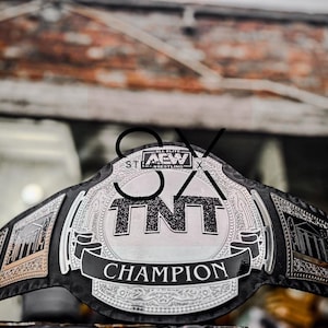 May include: A championship belt with a black leather strap and silver-colored metal accents. The belt features the text "TNT CHAMPION" on a black banner, with the letters "TNT" prominently displayed in the center. The words "AEW ALL ELITE WRESTLING" are also visible.