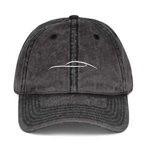 May include: A charcoal grey baseball cap with a distressed, washed-out finish. The front panel features a simple, white embroidered line drawing of a sleek car silhouette. The cap has a curved brim, a button on top, and stitched eyelets.