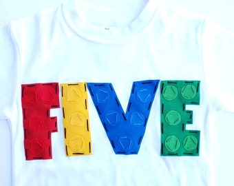 May include: White t-shirt with the word "FIVE" spelled out in colorful felt blocks that resemble Lego bricks. The letters are red, yellow, blue, and green.