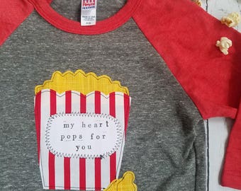 May include: A gray and red baseball tee shirt with a popcorn box design and the text "my heart pops for you".
