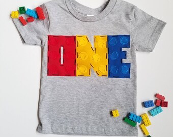 May include: A gray t-shirt with the word "ONE" spelled out in red, yellow, and blue Lego blocks. The shirt is surrounded by scattered Lego blocks.