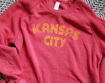 May include: Red sweatshirt with a yellow graphic that says "KANSAS CITY" in a retro font.