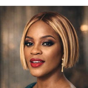 May include: A woman with a bob hairstyle, featuring blonde highlights, and wearing a teal patterned top. She has dark eye makeup and red lipstick. Gold earrings dangle from her ears. The background is blurred.