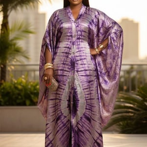 Puede incluir: Vestido largo morado y plateado con efecto tie-dye, con botones delanteros y mangas anchas. El vestido está adornado con pulseras doradas y un pequeño clutch dorado. La tela tiene un brillo sedoso.