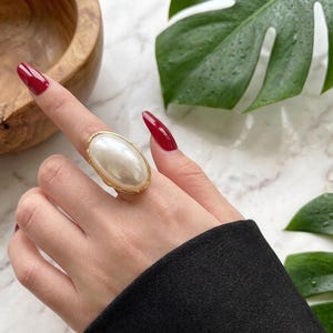 May include: A large, oval-shaped ring with a faux pearl center and a gold-colored metal frame. The ring is worn on a finger, with a black sleeve visible in the image. The background includes a wooden bowl and green leaves.